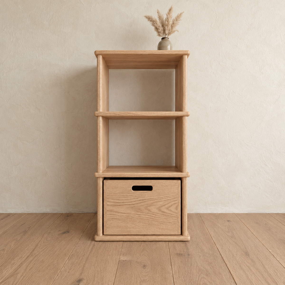 The small bookshelf in white oak against a plaster wall, with a small vase of dried grass on top and a wooden bin on the bottom shelf.