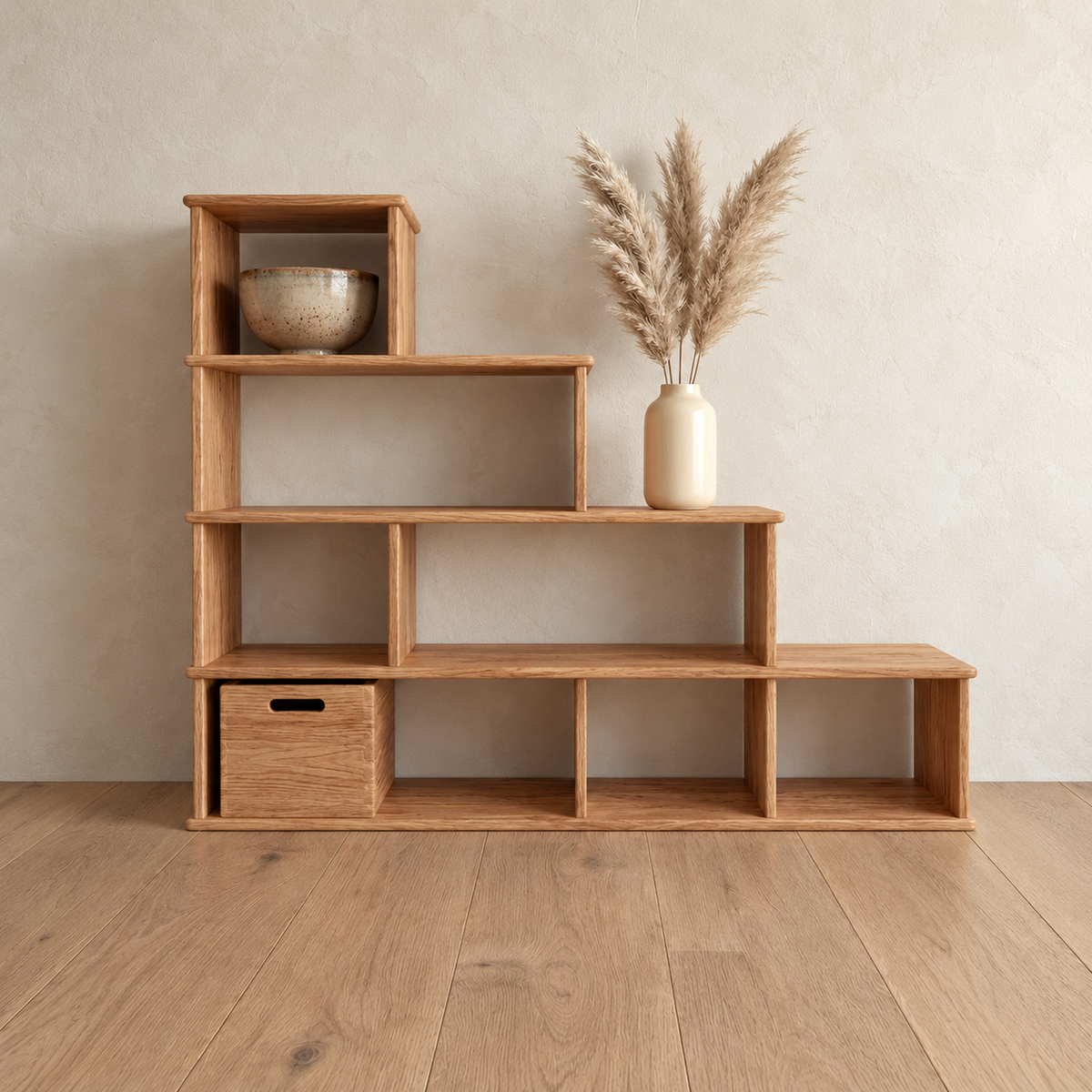 The step bookshelf in red oak against a plaster wall, with a bowl in the top cube, a vase of pampas grass on a step and a wooden storage cube in the bottom row.