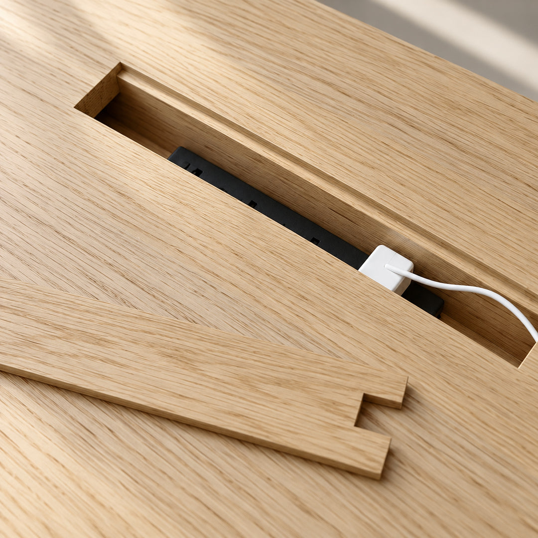 The conference table in white oak with its lid lifted out, showing the black power bar in the channel below and a charger plugged in.