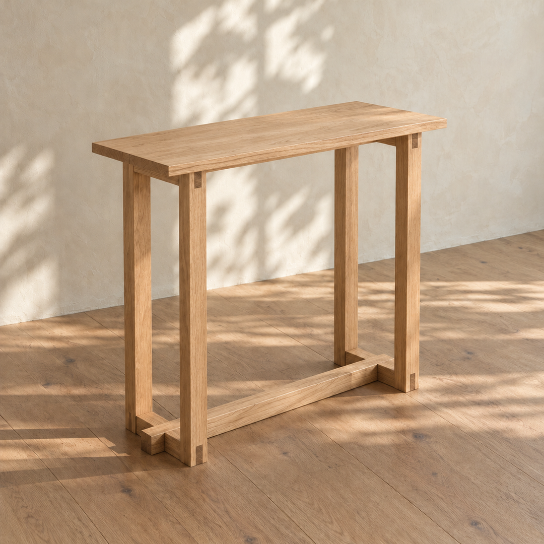 The 2 person bar table in white oak from an angle in dappled sunlight, with its solid top, bridle-jointed leg frames and a long stretcher near the floor.