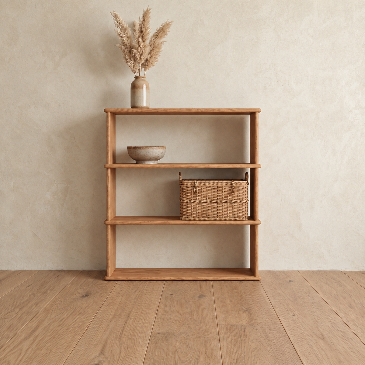 The 3 shelf bookcase in red oak against a plaster wall, with a vase of pampas grass on top, a ceramic bowl on the upper shelf and a woven basket on the lower shelf.