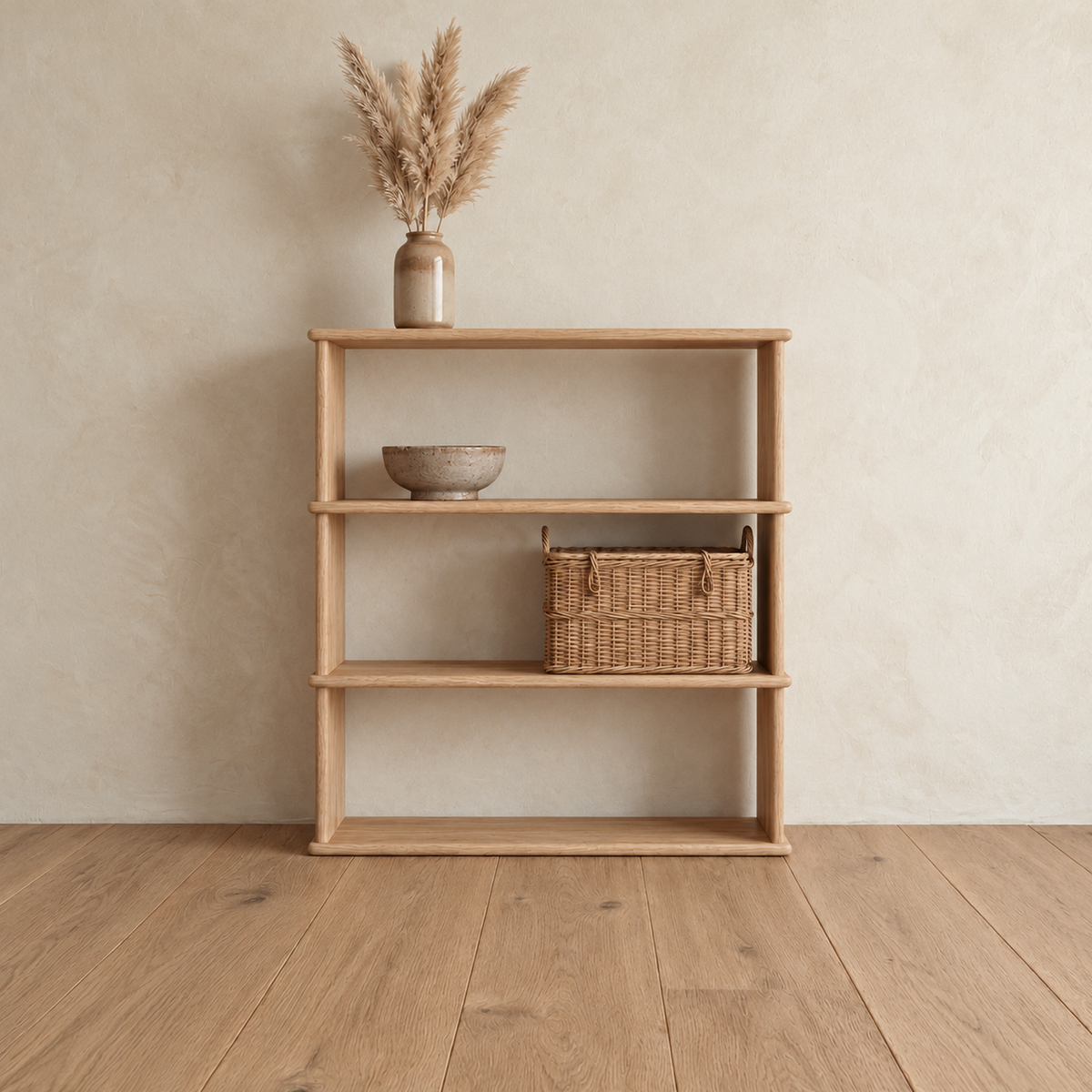 The 3 shelf bookcase in white oak against a plaster wall, with a vase of pampas grass on top, a ceramic bowl on the upper shelf and a woven basket on the lower shelf.