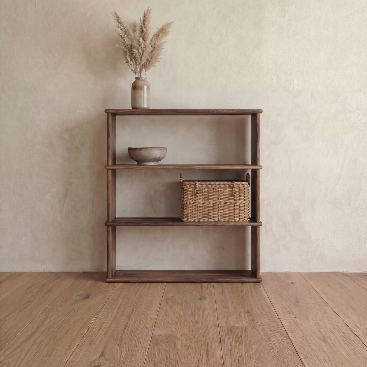 A 3 shelf bookcase in walnut against a plaster wall, with a vase of pampas grass on top and a bowl and woven basket on the shelves.
