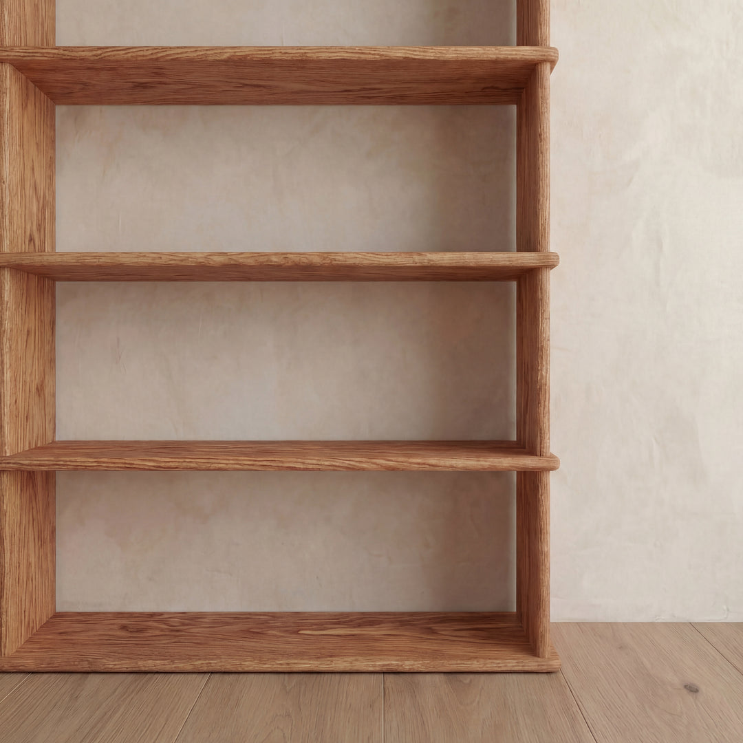 A 5 shelf bookcase in red oak with four visible shelves stands against a beige textured wall on a wooden floor, positioned on the left side of the image.