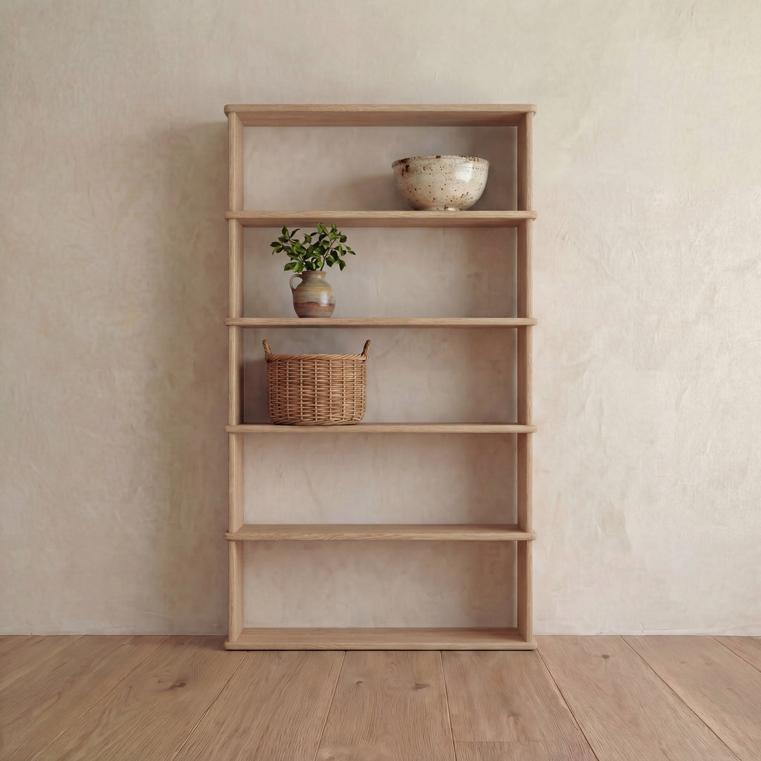 A 5 shelf bookcase in white oak stands against a light textured wall, displaying a ceramic bowl, a small potted plant, and a woven basket. The remaining shelves are empty, and the room features a wooden floor.
