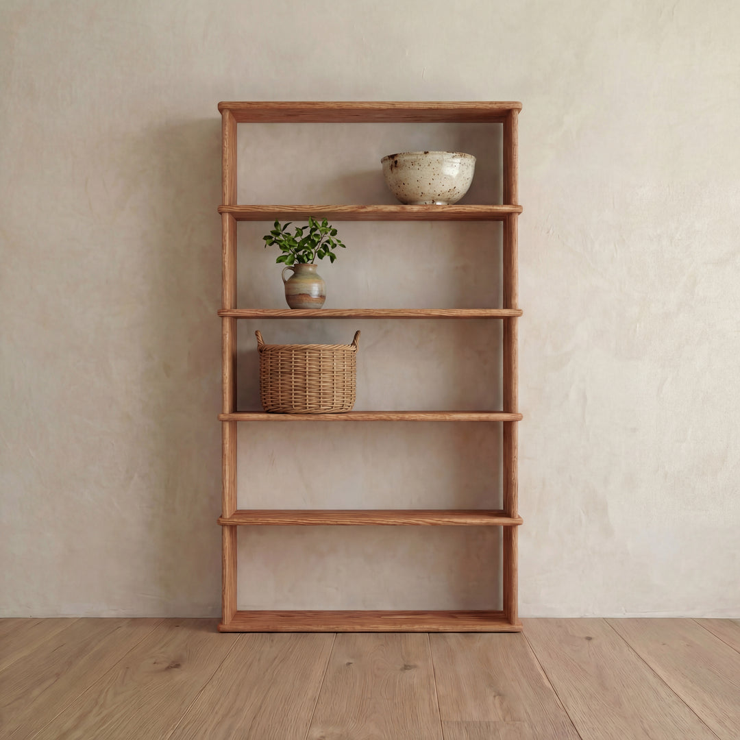 A 5 shelf bookcase in red oak stands against a light beige wall, displaying a ceramic bowl, a potted plant, and a woven basket on its minimalist handmade shelves, with most left empty.