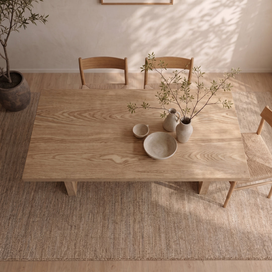 A 6 person dining table in white oak is paired with three chairs, minimalist ceramic dishes, and a vase filled with leafy branches, all arranged on a woven rug in a softly lit Vancouver room featuring a potted plant in the corner.