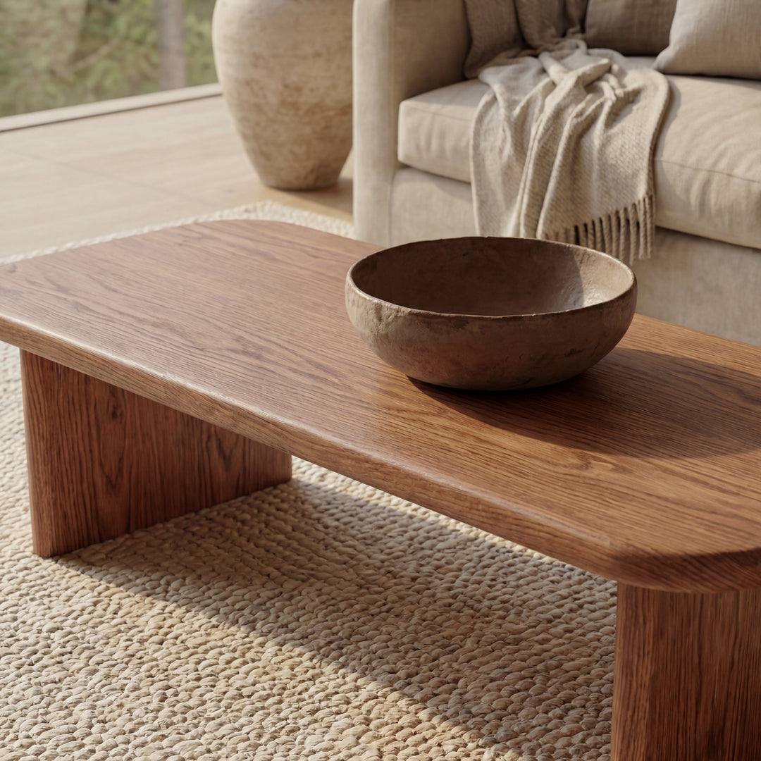 A 60 inch coffee table in red oak is showcased on a textured woven rug in a cozy living room, with a beige sofa and draped blanket softly lit by natural light in the background.