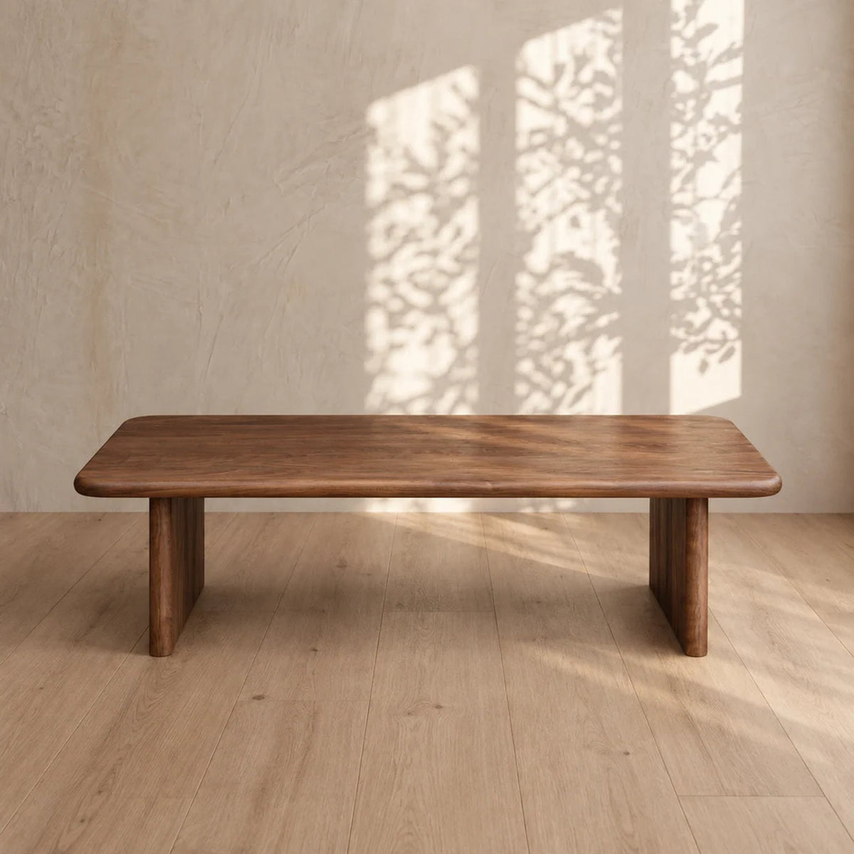 A 60 inch coffee table in walnut on a light wood floor, with leafy window shadows on the plaster wall behind it.