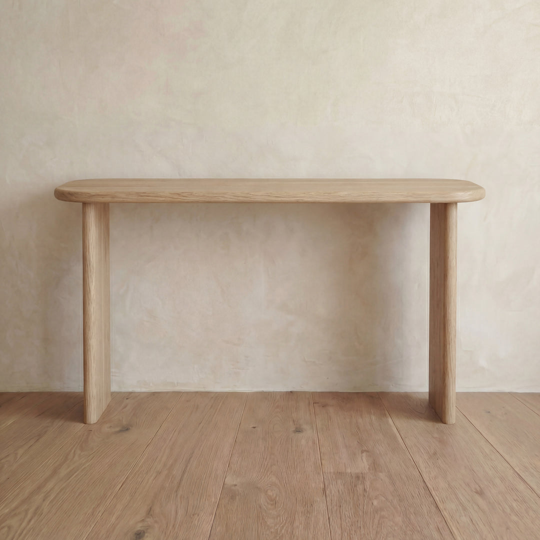 A 60 inch console table in white oak with rounded edges and three legs stands on a wooden floor against a neutral, textured wall.