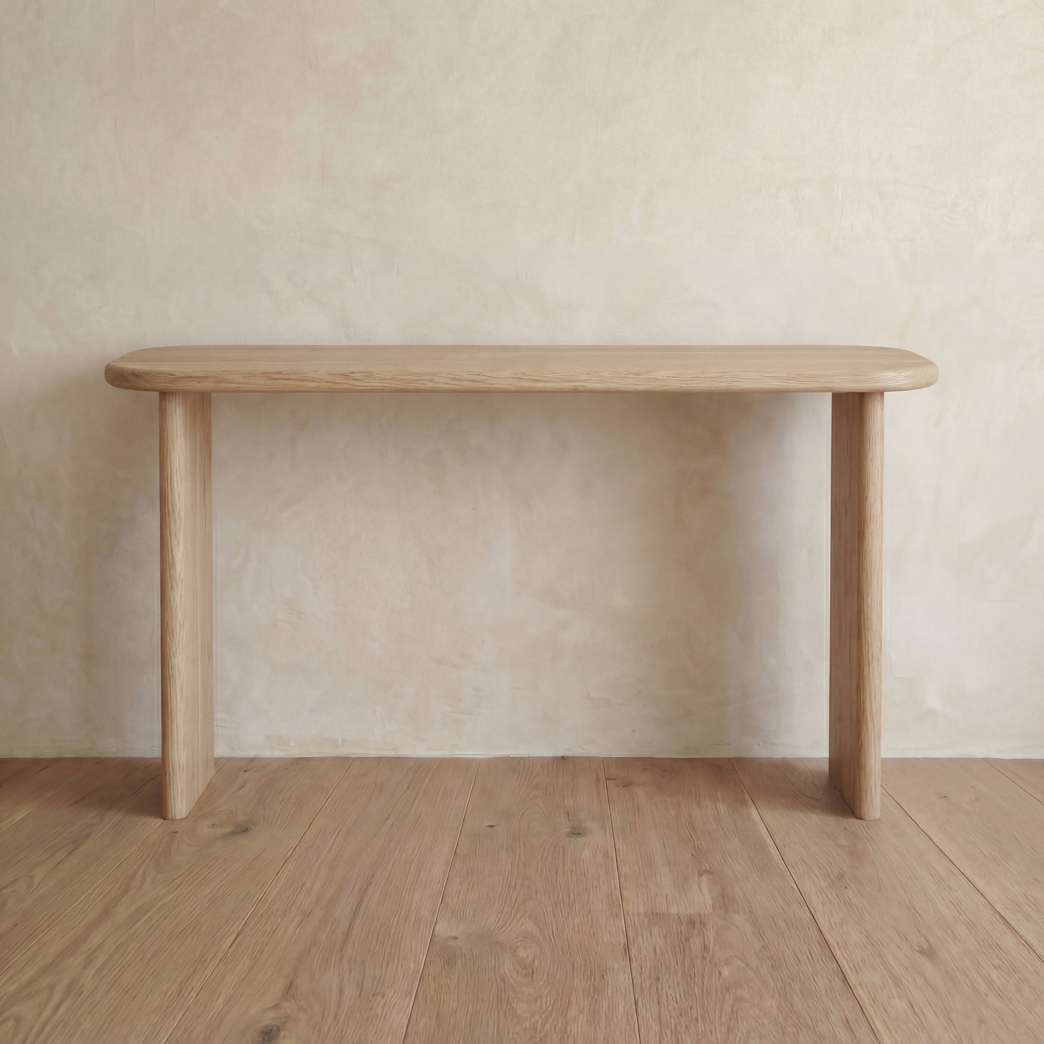 A 60 inch console table in white oak with rounded edges and three legs stands on a wooden floor against a neutral, textured wall.