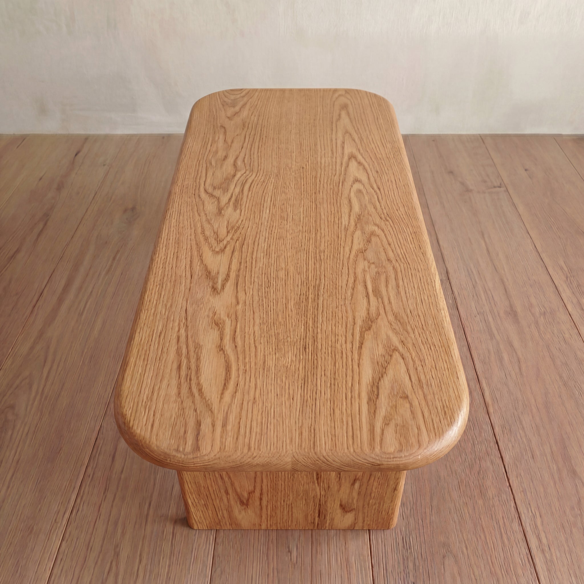 The 60 inch coffee table in red oak, handmade with rounded edges and visible wood grain, is placed on a matching wooden floor next to a light-colored wall.