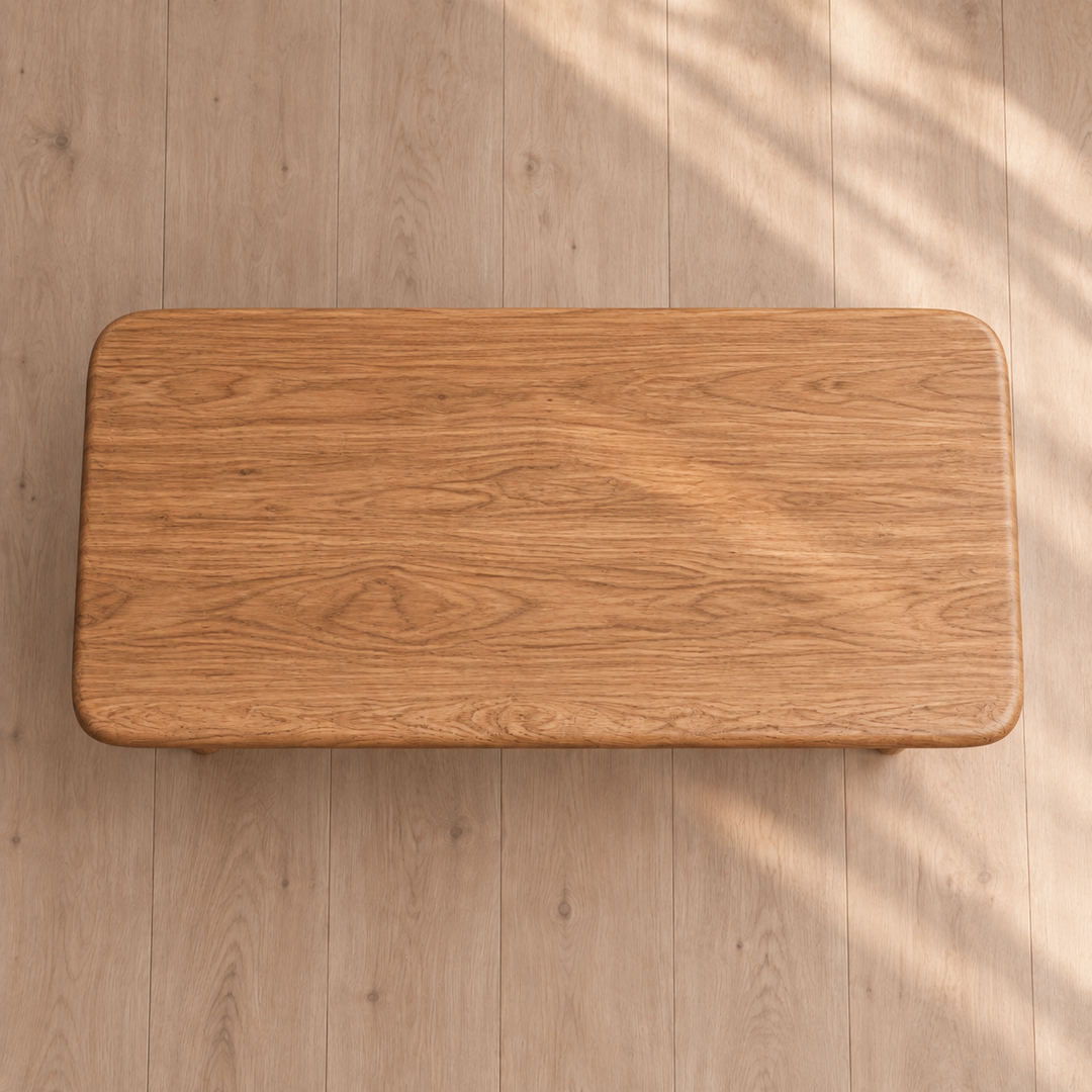 A 60 inch coffee table in red oak with rounded corners sits on a light wood floor, illuminated by natural sunlight that casts soft shadows across the minimalist scene.
