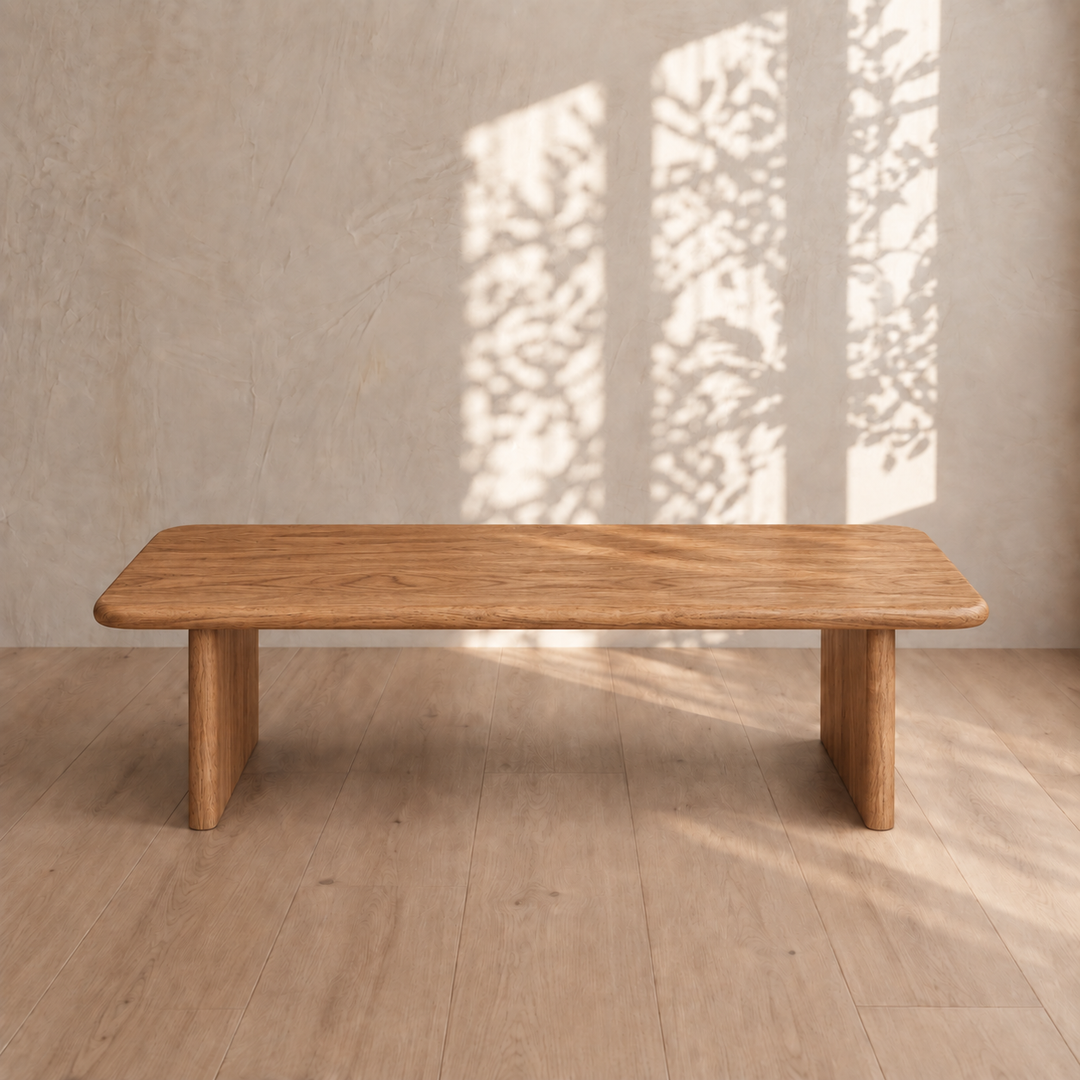A 60 inch coffee table in red oak sits on a light wood floor. Sunlight pours in from the right, casting leaf-shaped shadows across a beige wall behind it. The scene exudes a minimalist, natural aesthetic.