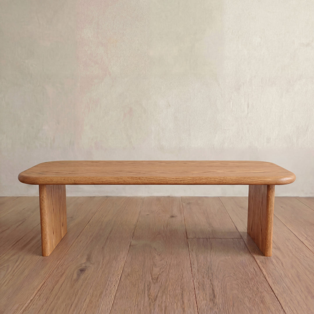 A sleek, modern 60 inch coffee table in solid red oak with rounded edges and rectangular legs sits on a wooden floor against a light-colored wall.