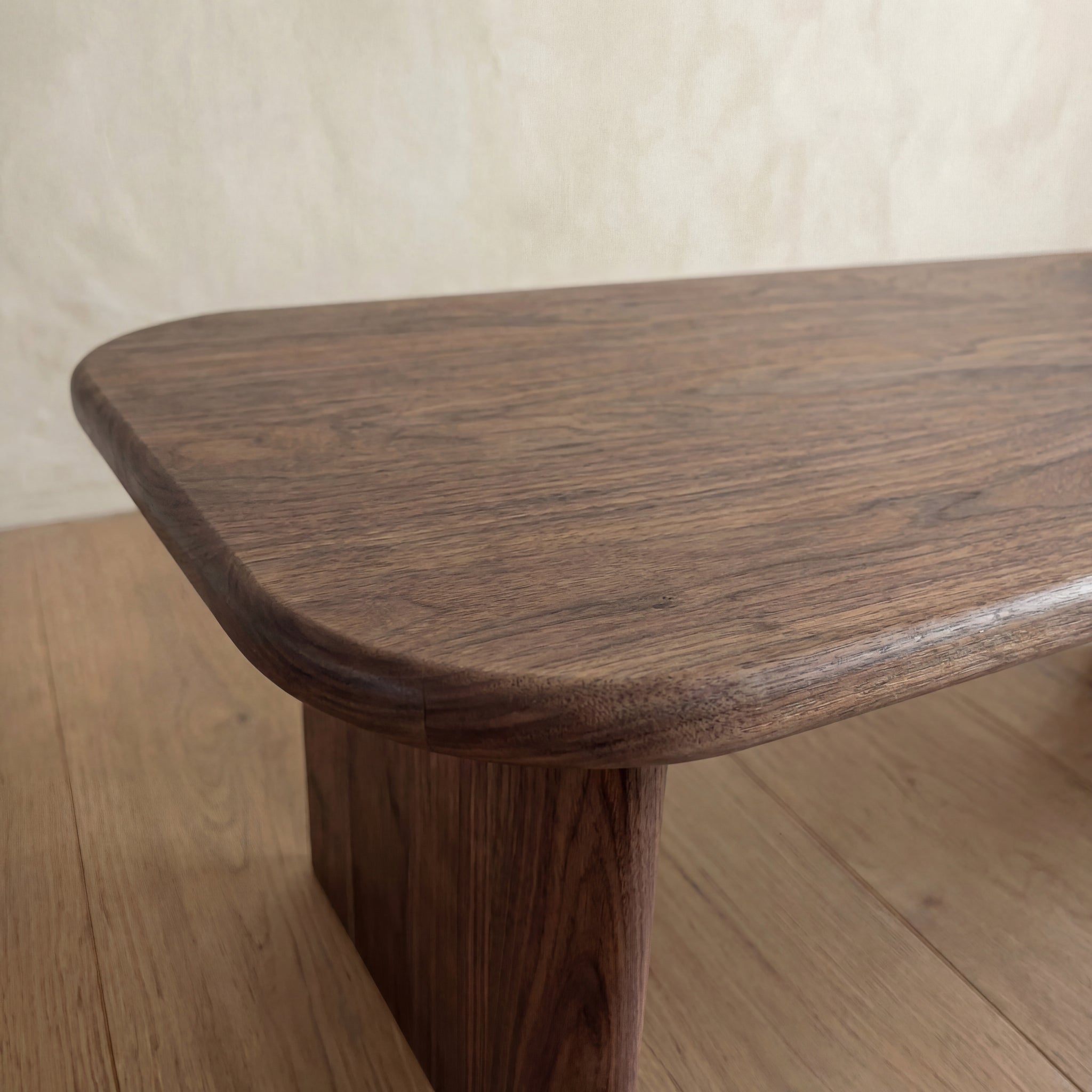 Close-up of a 60 inch coffee table in walnut with rounded edges and a smooth solid surface, placed on a light wood floor against a beige wall background.