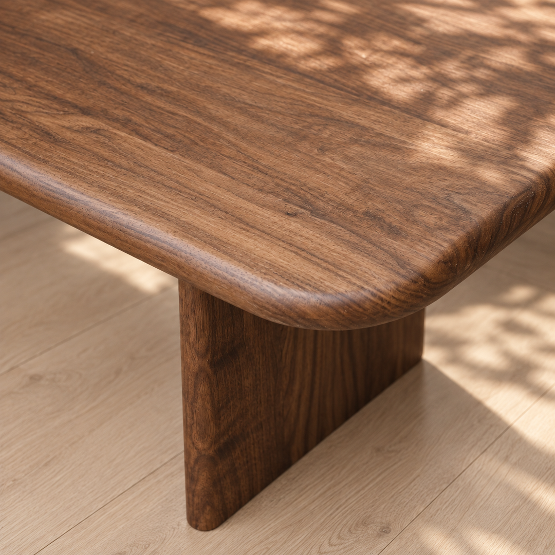 Close-up of a handmade 60 inch coffee table in walnut with rounded edges, set on a light wood floor. Sunlight casts soft, leaf-patterned shadows across the table’s smooth, solid walnut surface.