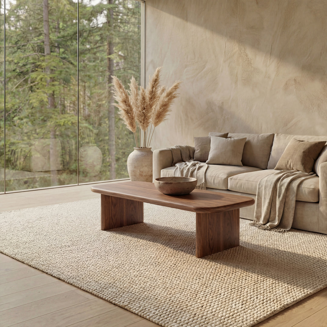 A modern living room with neutral tones includes a beige sofa, a 60 inch coffee table in walnut, a large vase holding pampas grass, a textured rug, and floor-to-ceiling windows revealing views of the forest.