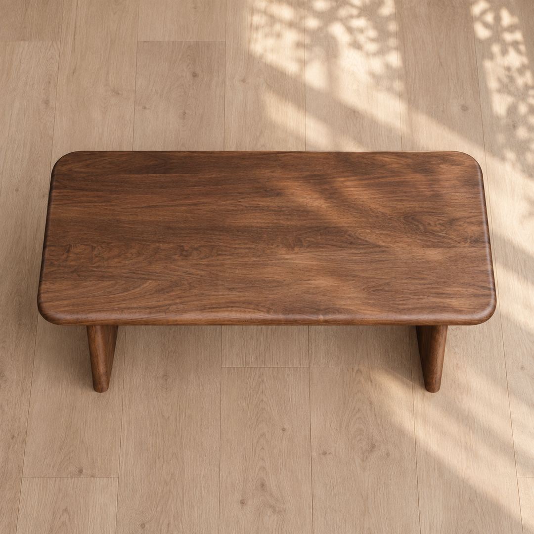A 60 inch coffee table in walnut with a rectangular handmade design and rounded edges sits on a light wood floor. Sunlight and leafy shadows play across the floor, enhancing the natural ambiance of the scene.