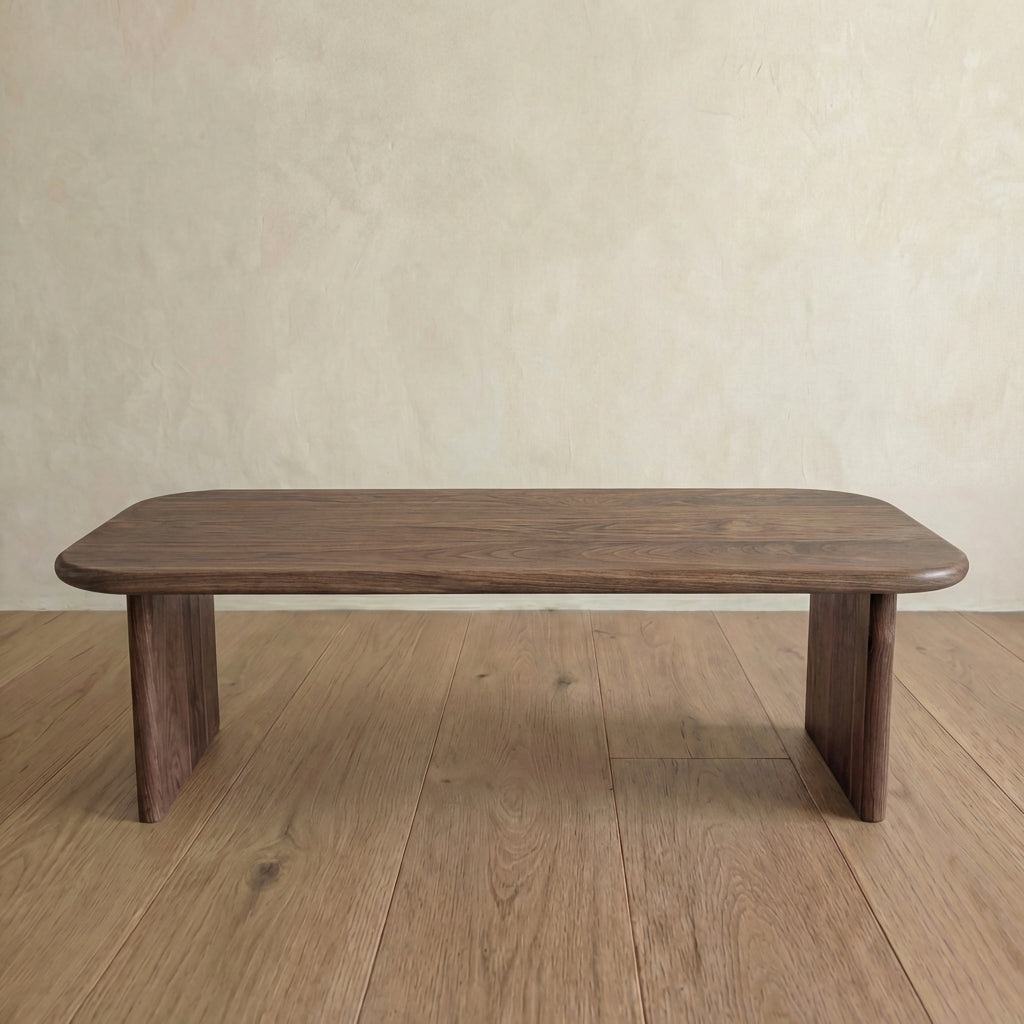 The 60 inch coffee table in walnut features a minimalist, handmade design with rounded edges and two wide legs, placed on a wooden floor against a light beige textured wall.