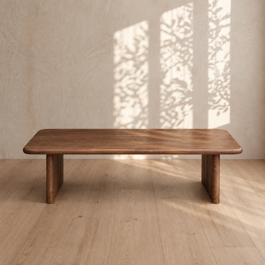 A 60 inch coffee table in walnut sits on a light wood floor, while sunlight pours through a window, casting leaf-shaped shadows on the beige wall behind it.