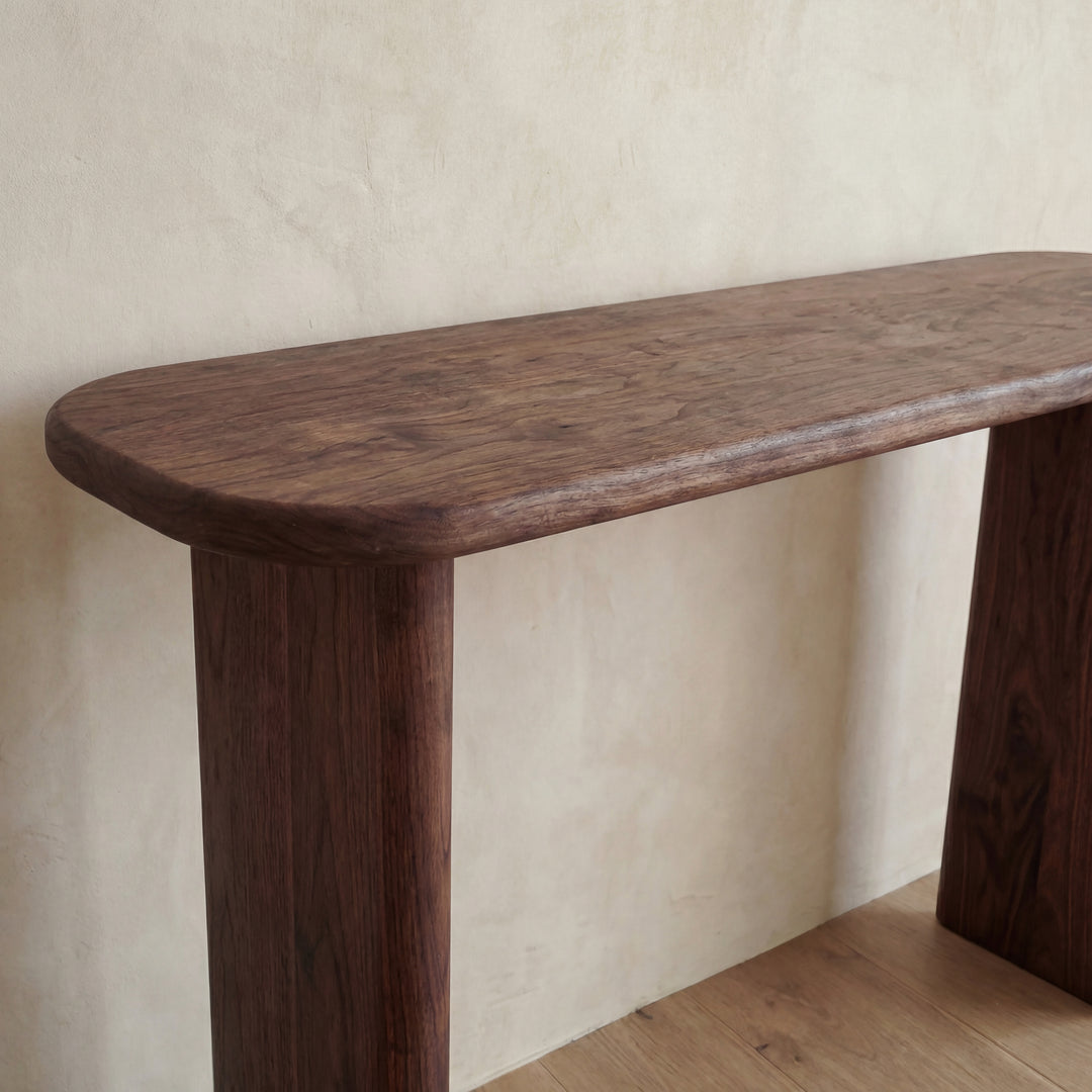 The 60 inch console table in walnut features a minimalist design with rounded edges and a smooth finish, standing against a light beige textured wall on a wooden floor. Handmade in Vancouver.
