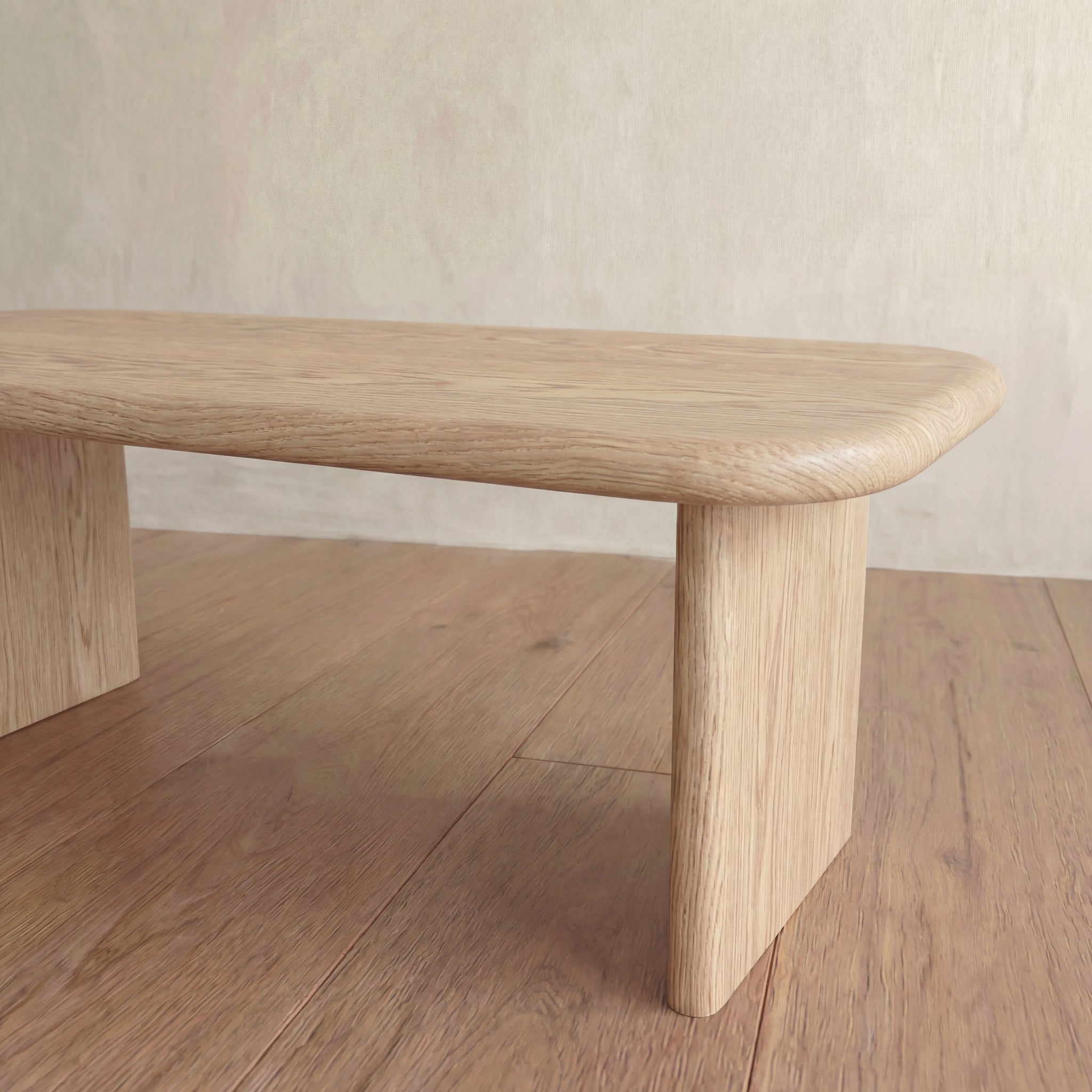 A 60 inch coffee table in white oak with rounded edges and sturdy legs rests on a wooden floor against a pale, textured wall.