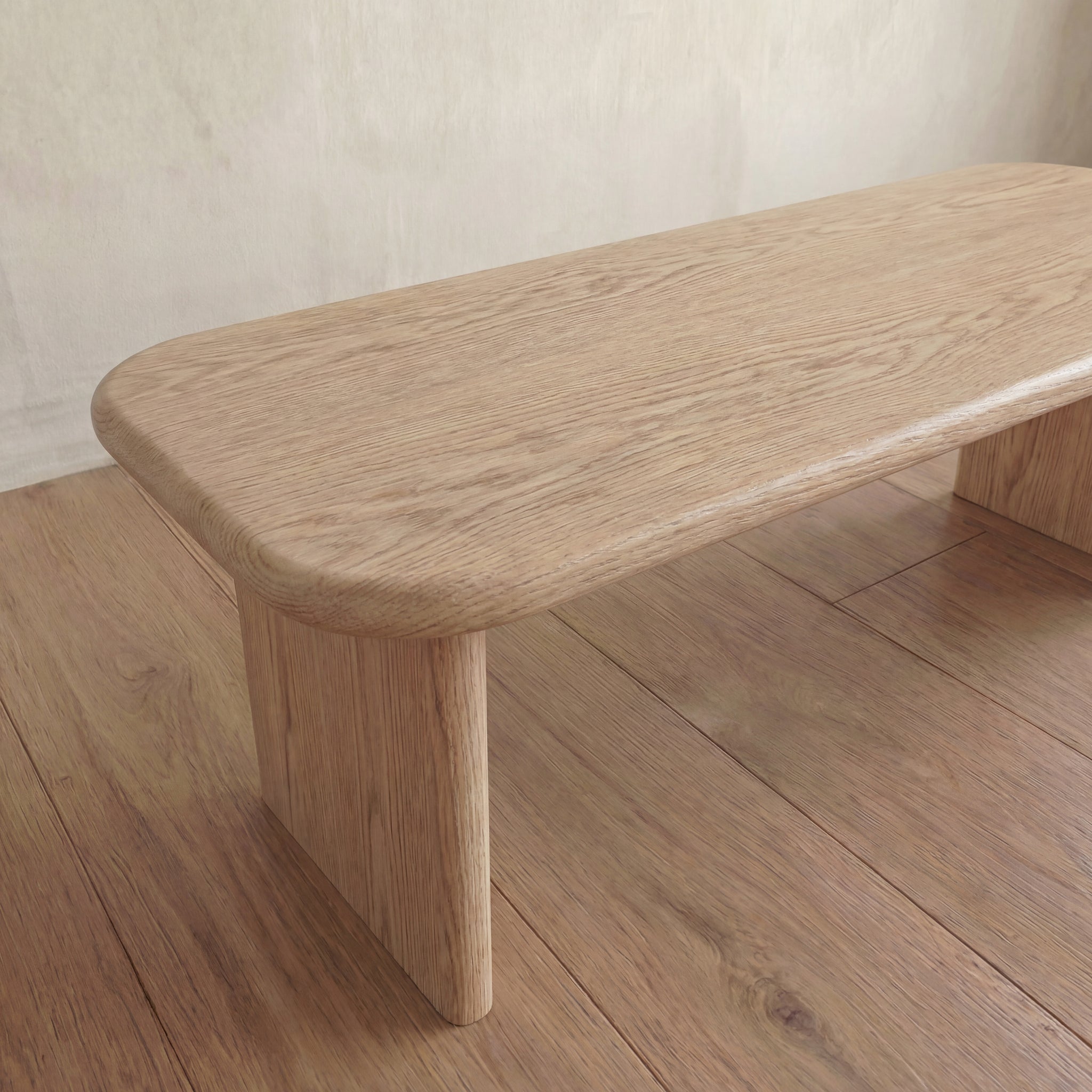 A 60 inch white oak coffee table with rounded edges and a light natural finish sits on a matching wooden floor against a neutral-colored wall.