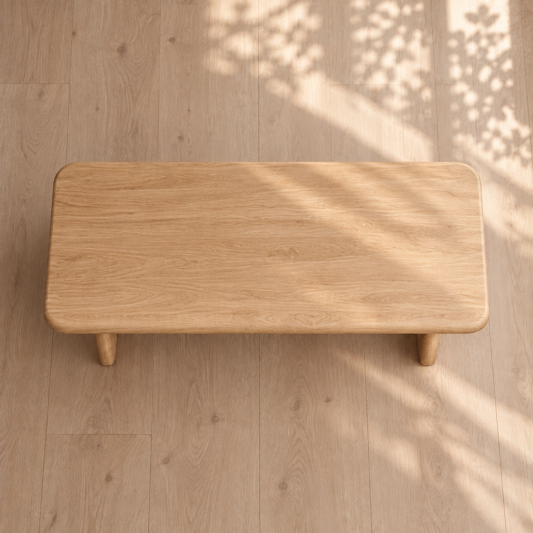 A 60 inch coffee table in white oak with a rectangular shape and rounded corners rests on a matching wooden floor. Sunlight filters through leaves, casting delicate shadows across the surface and floor, emphasizing the elegant simplicity of this minimalist coffee table.