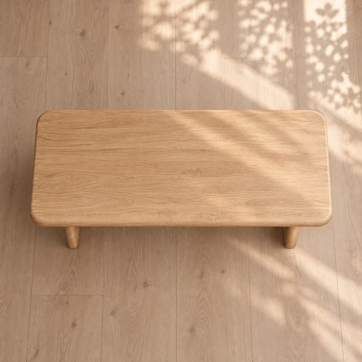A 60 inch coffee table in white oak with a rectangular shape and rounded corners rests on a matching wooden floor. Sunlight filters through leaves, casting delicate shadows across the surface and floor, emphasizing the elegant simplicity of this minimalist coffee table.