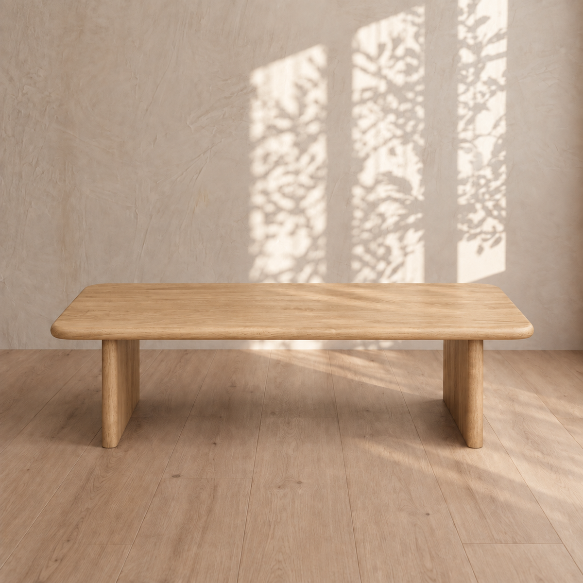 A 60 inch coffee table in white oak adds a touch of elegance to a sunlit room with wooden floors and textured beige walls. Leaf shadows dance on the wall, enhancing the calming and natural ambiance around this minimalist centerpiece.