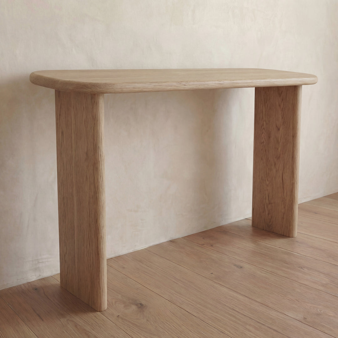 The 60 inch console table in white oak, featuring rounded edges and thick legs, stands against a light beige wall. Its solid build and natural wood grain finish complement the wooden floor’s similar texture.