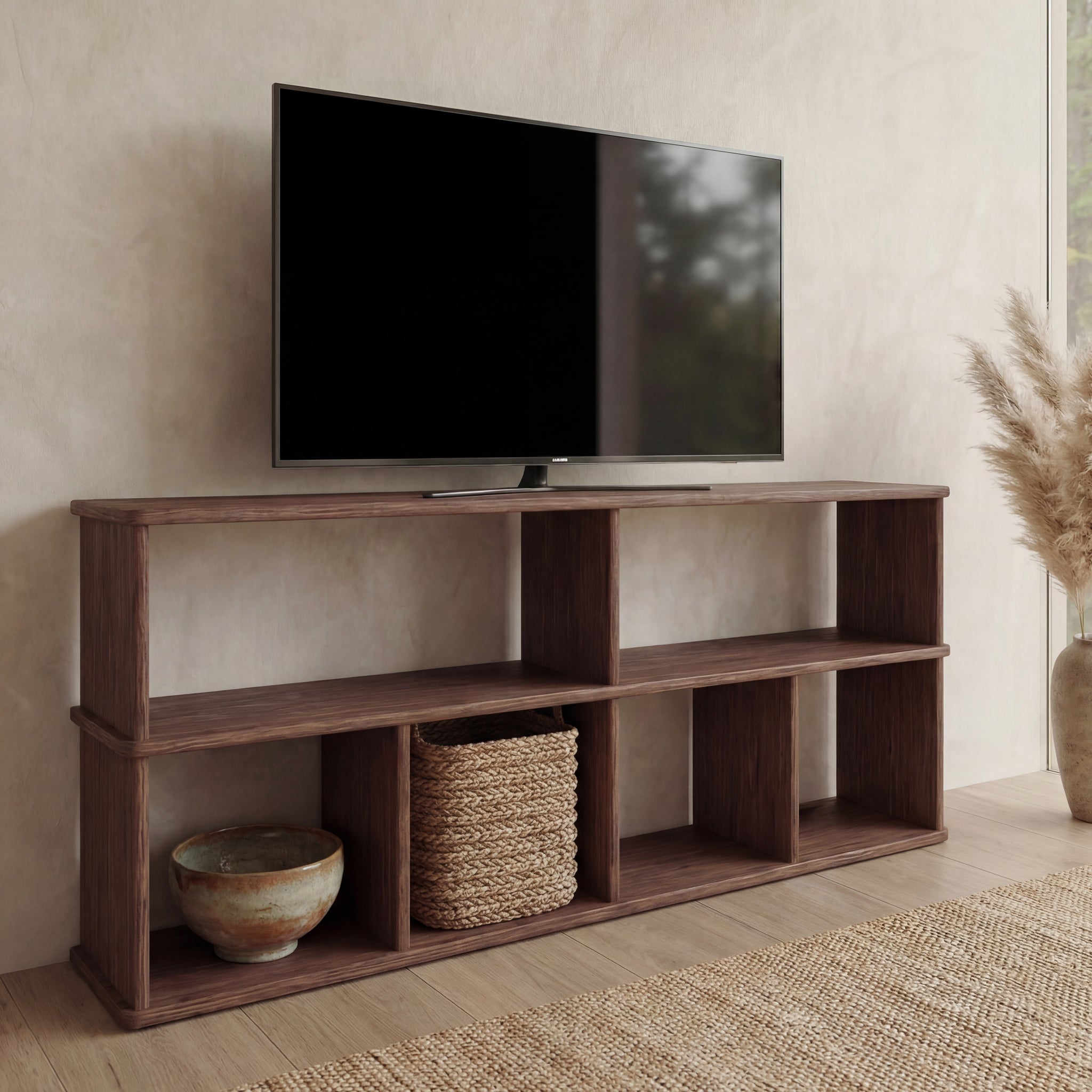 A 65 inch flat-screen TV is displayed on a walnut bookshelf TV stand with open shelves, which hold a woven basket and ceramic bowl. Nearby, pampas grass decorates the light wood floor.