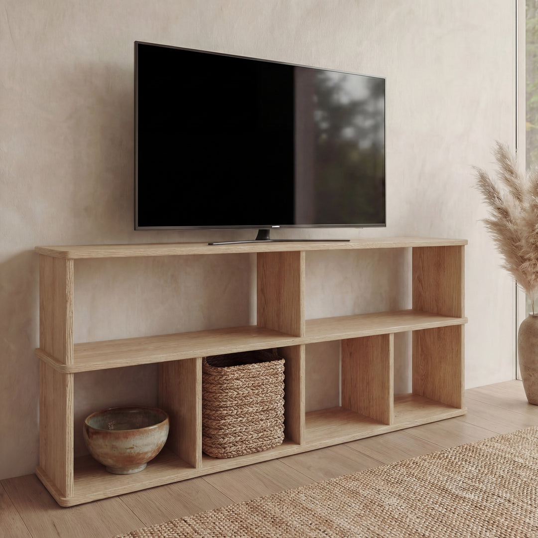 A 65 inch bookshelf TV stand in white oak with open shelves displays a flat-screen TV, woven basket, ceramic bowl, and pampas grass in a vase, set against neutral-toned walls and flooring.
