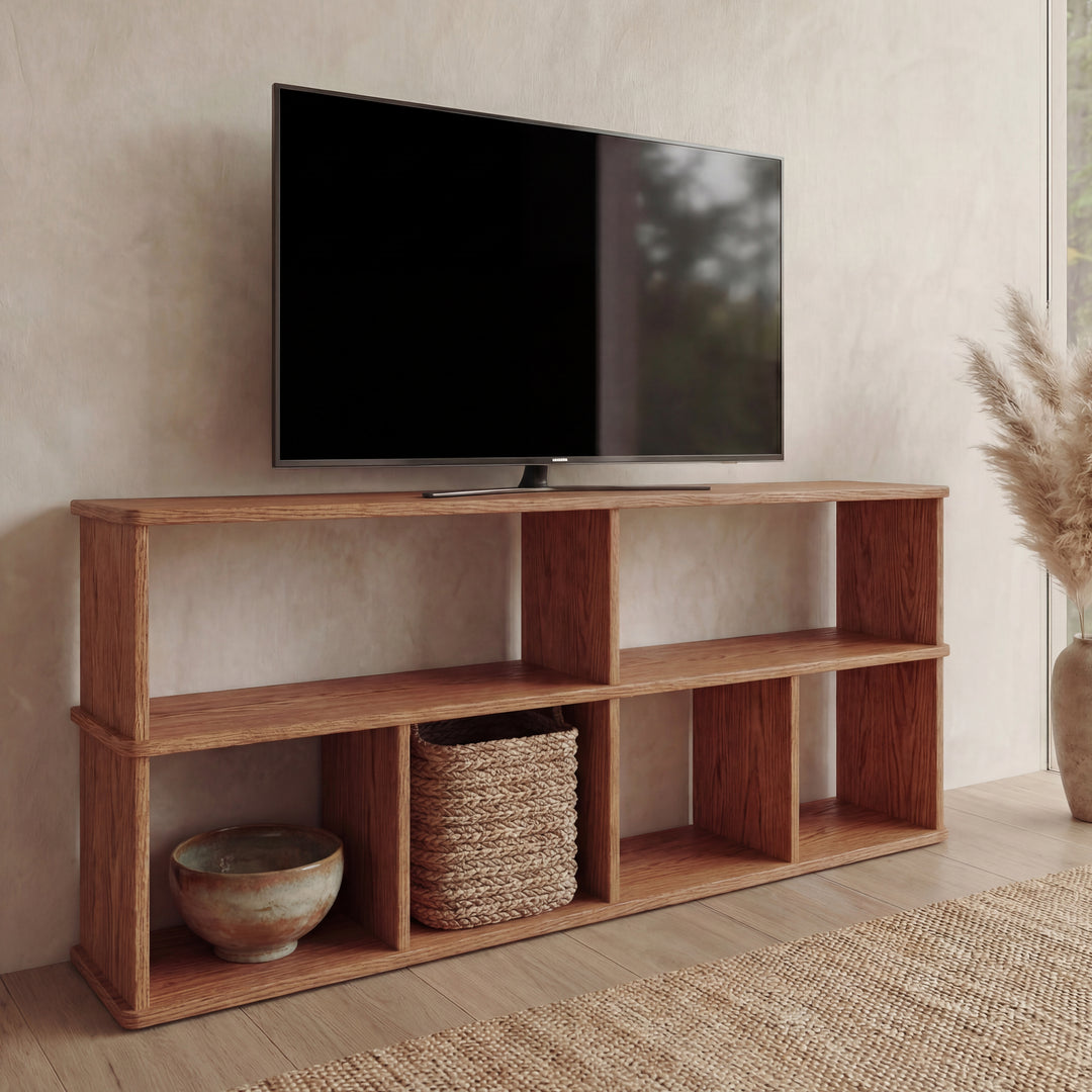 A flat-screen TV is on a 65 inch bookshelf TV stand in red oak with open shelves, which display a woven basket and ceramic bowl. Pampas grass in a vase and a woven rug are on the floor, sunlight streaming in from the right.