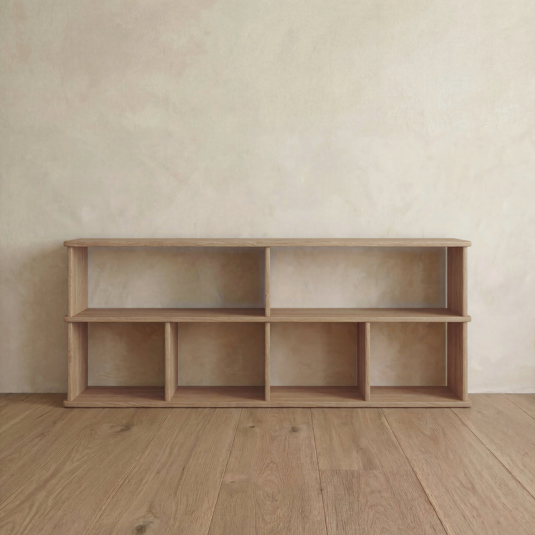 The 65 inch bookshelf TV stand in white oak stands on a wooden floor by a light beige textured wall. It features six rectangular compartments, clean lines, and a minimalist design with a natural wood finish.