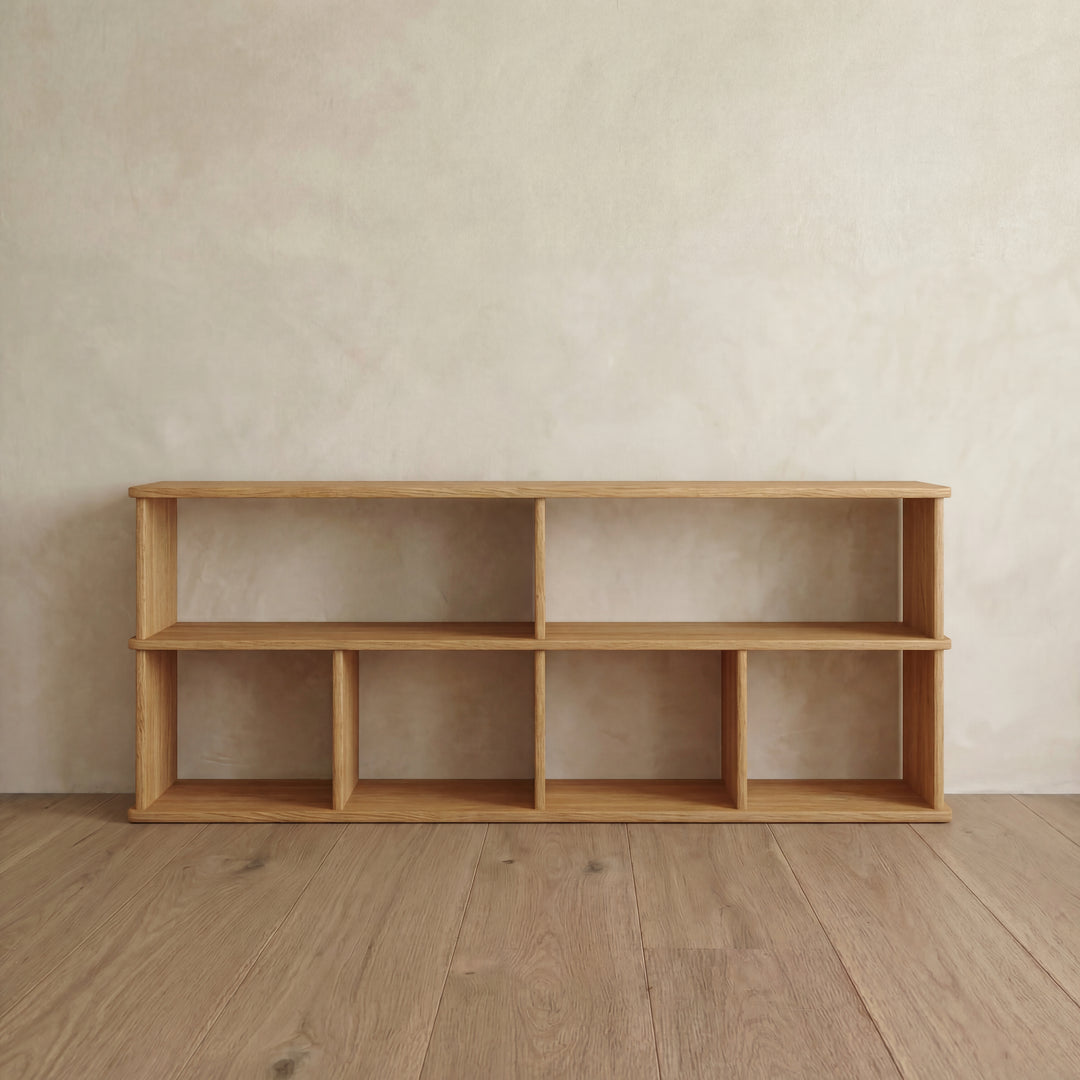 The 65 inch bookshelf TV stand in white oak stands on a wooden floor by a light beige textured wall. It features six rectangular compartments, clean lines, and a minimalist design with a natural wood finish.