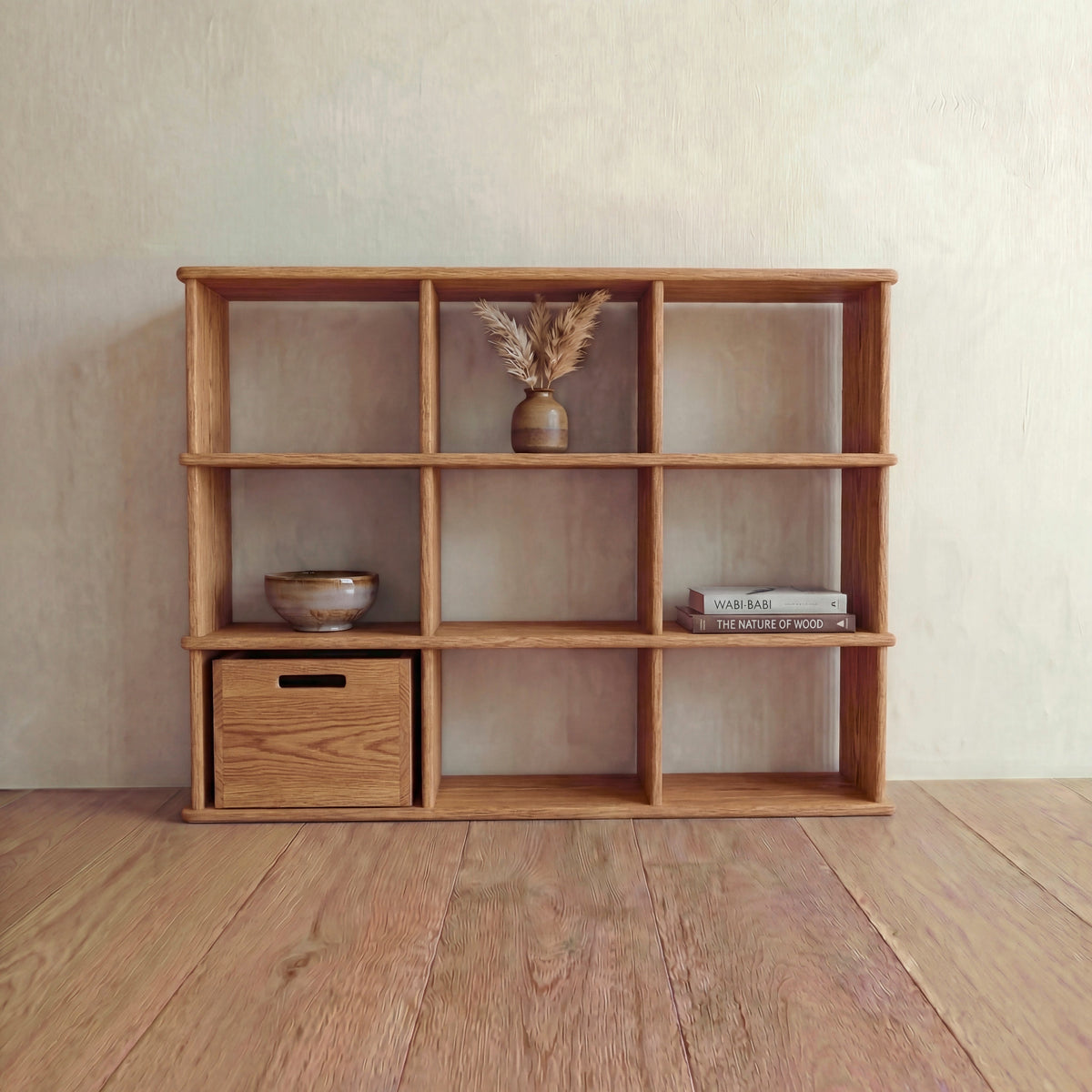 The Nine cube bookshelf in red oak, its nine square compartments holding a bowl, a storage cube, a vase of dried grasses and stacked books, against a light wall on a wooden floor.