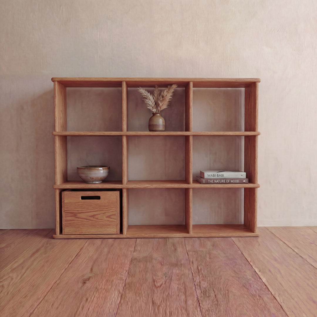 The Nine cube bookshelf in red oak features nine square compartments displaying a bowl, small drawer, vase with dried grasses, and stacked books. It stands against a light-colored wall on a wooden floor.