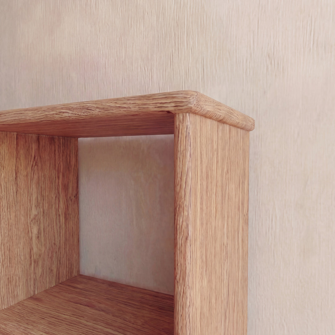A close-up of the top corner of the Nine cube bookshelf in red oak against a light, textured wall highlights its natural wood grain finish and simple, rectangular design.