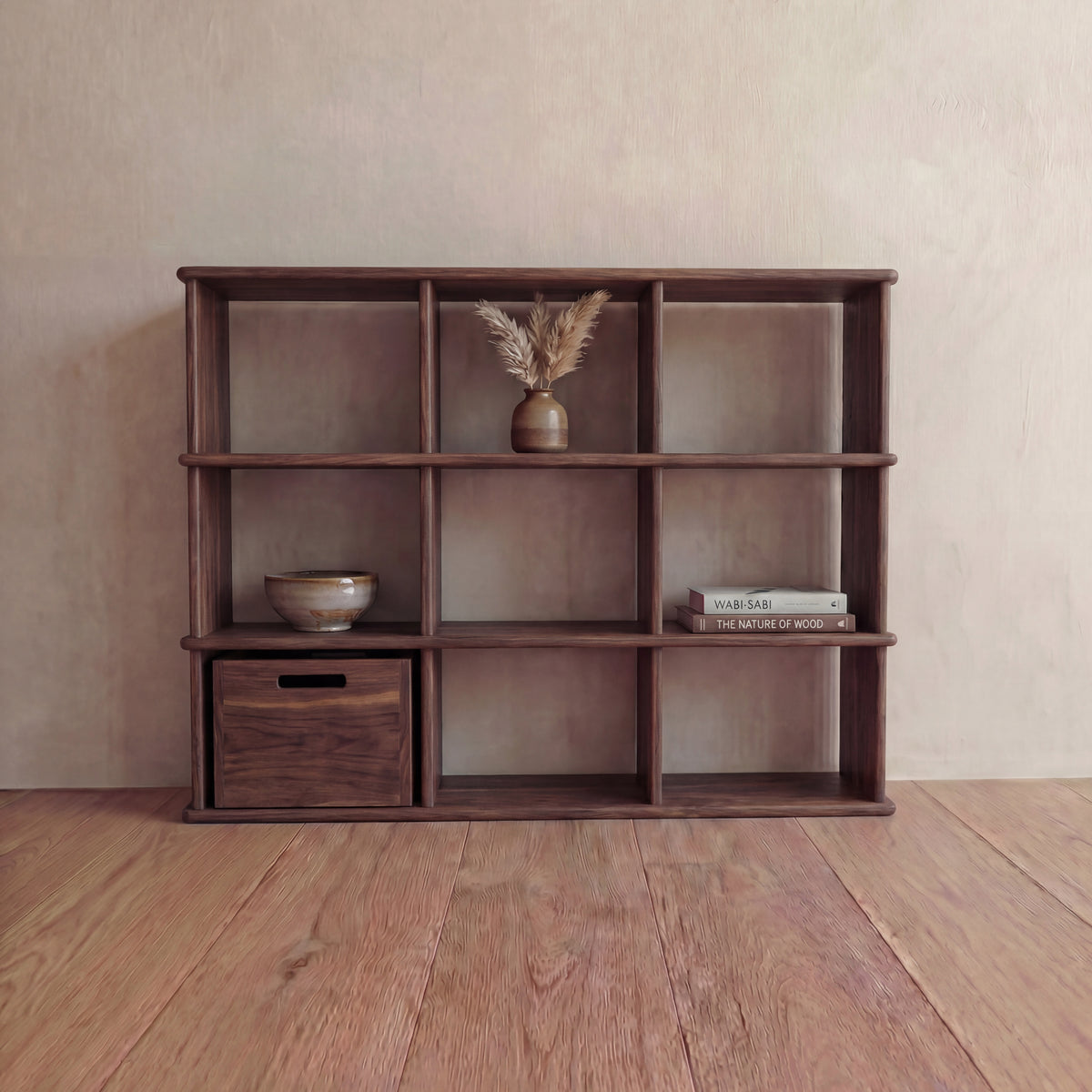 The Nine cube bookshelf in walnut, handmade in Vancouver, displays a small vase with dried grass, a bowl, a wooden box, and two books against a light-colored wall and wooden floor.