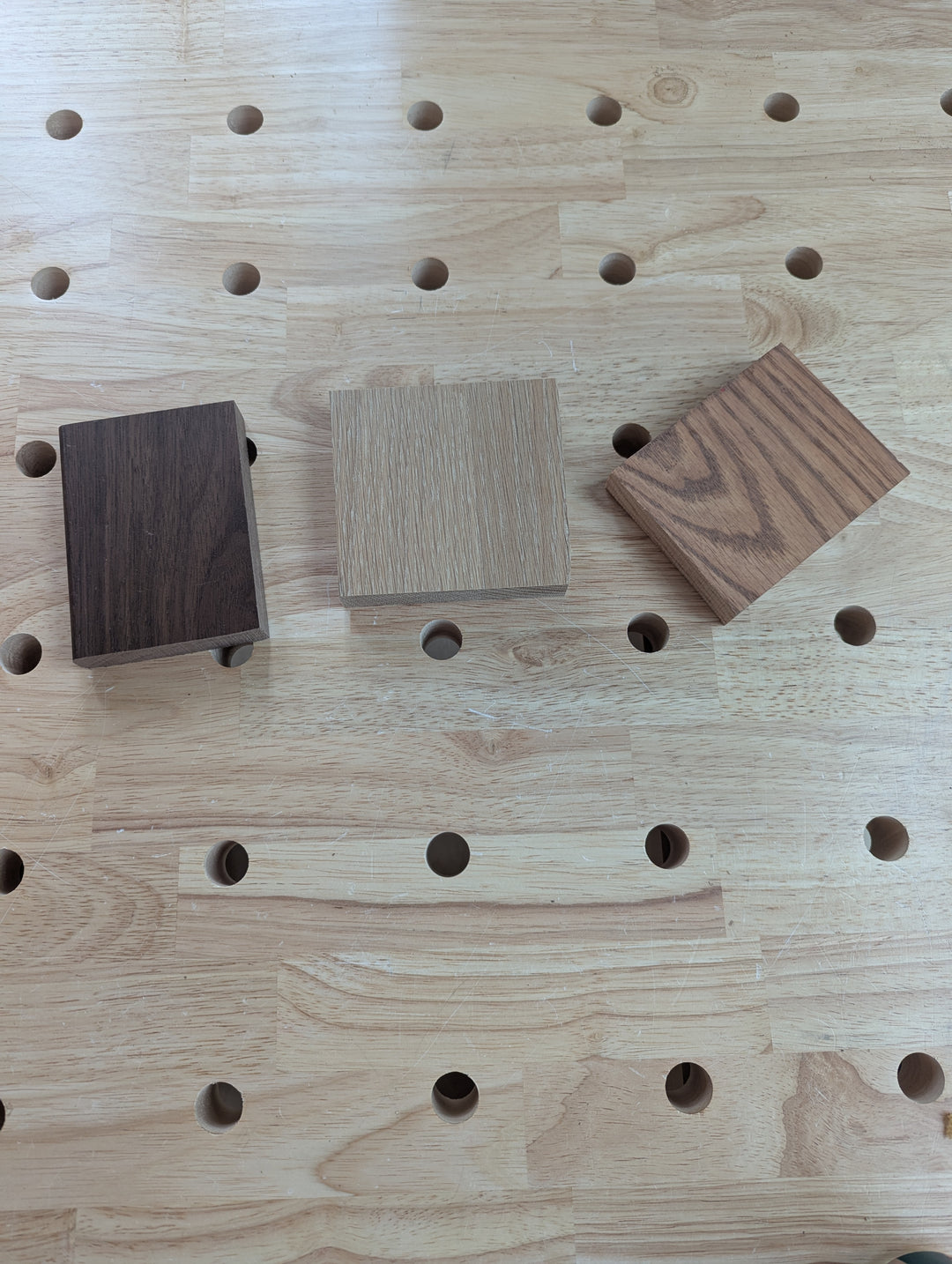 Three small rectangular Wood Samples—dark brown, light beige, and medium brown—are arranged side by side on a light workbench with evenly spaced holes. These Wood Samples are ready to ship within 2-3 days.