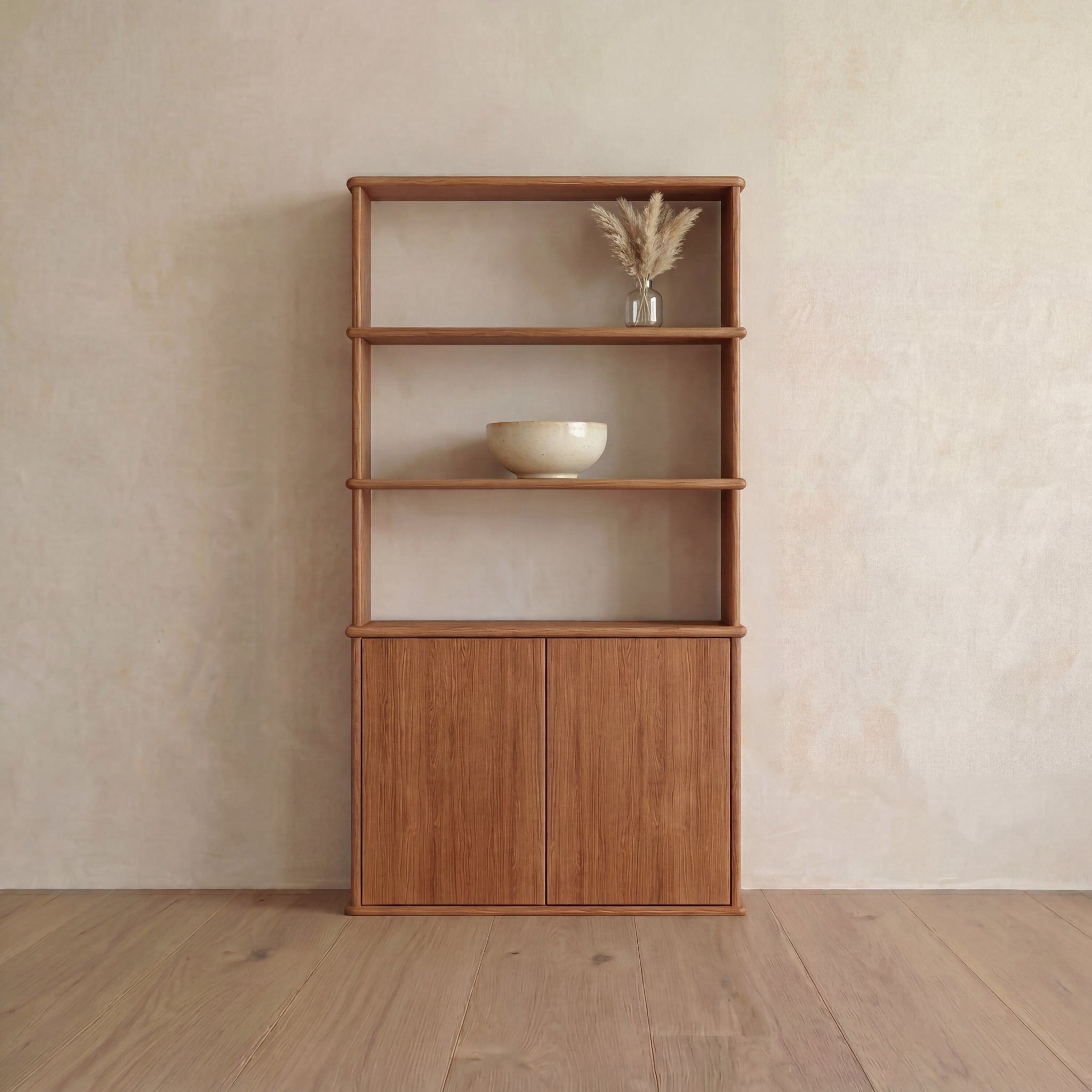 The Cabinet bookshelf in red oak, inspired by Vancouver, features push-to-open doors at the bottom. An off-white bowl sits on the middle shelf, while a glass vase with dried grasses is displayed on the top shelf. The bookshelf is set against a beige wall and wooden floor.
