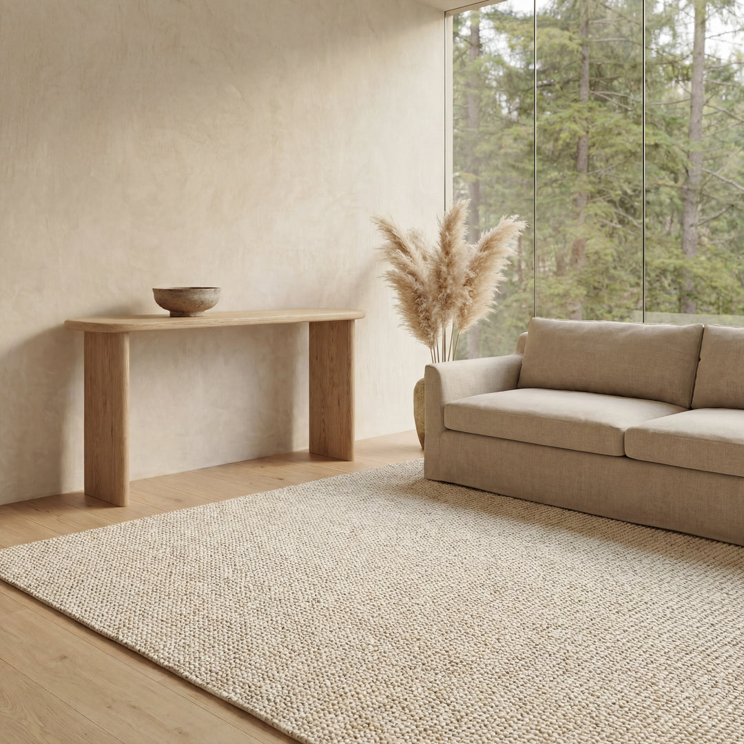 A minimalist living room features a light beige sofa, neutral rug, and a 60 inch console table in white oak topped with a bowl and pampas grass. Floor-to-ceiling windows showcase views of the lush green forest outside.
