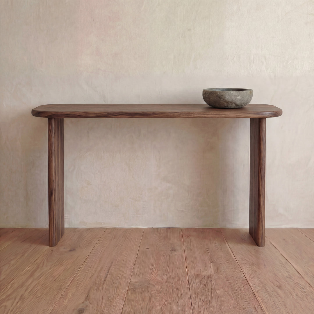 The 60 inch console table in walnut, handmade in Vancouver, stands on a wooden floor against a light textured wall. A rustic stone bowl on the right side adds to its understated elegance.