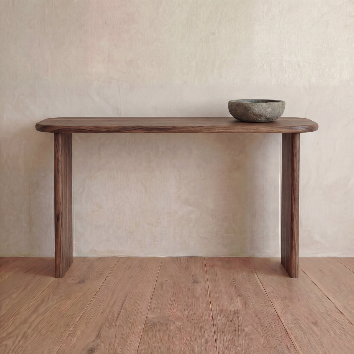 The 60 inch console table in walnut, handmade in Vancouver, stands on a wooden floor against a light textured wall. A rustic stone bowl on the right side adds to its understated elegance.