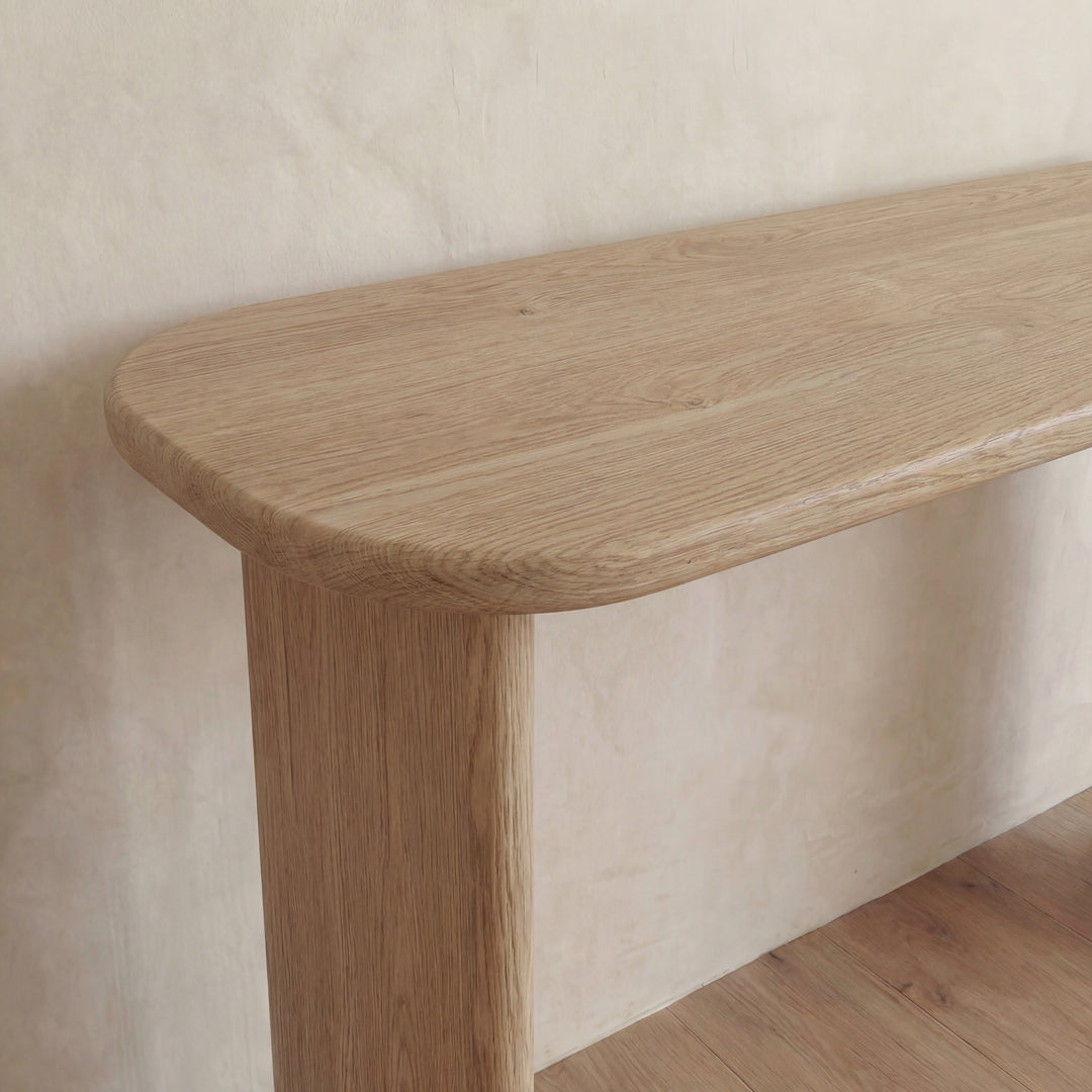 A close-up of the 60 inch console table in white oak, featuring rounded corners and a light natural finish, set against a textured beige wall on a wooden floor.