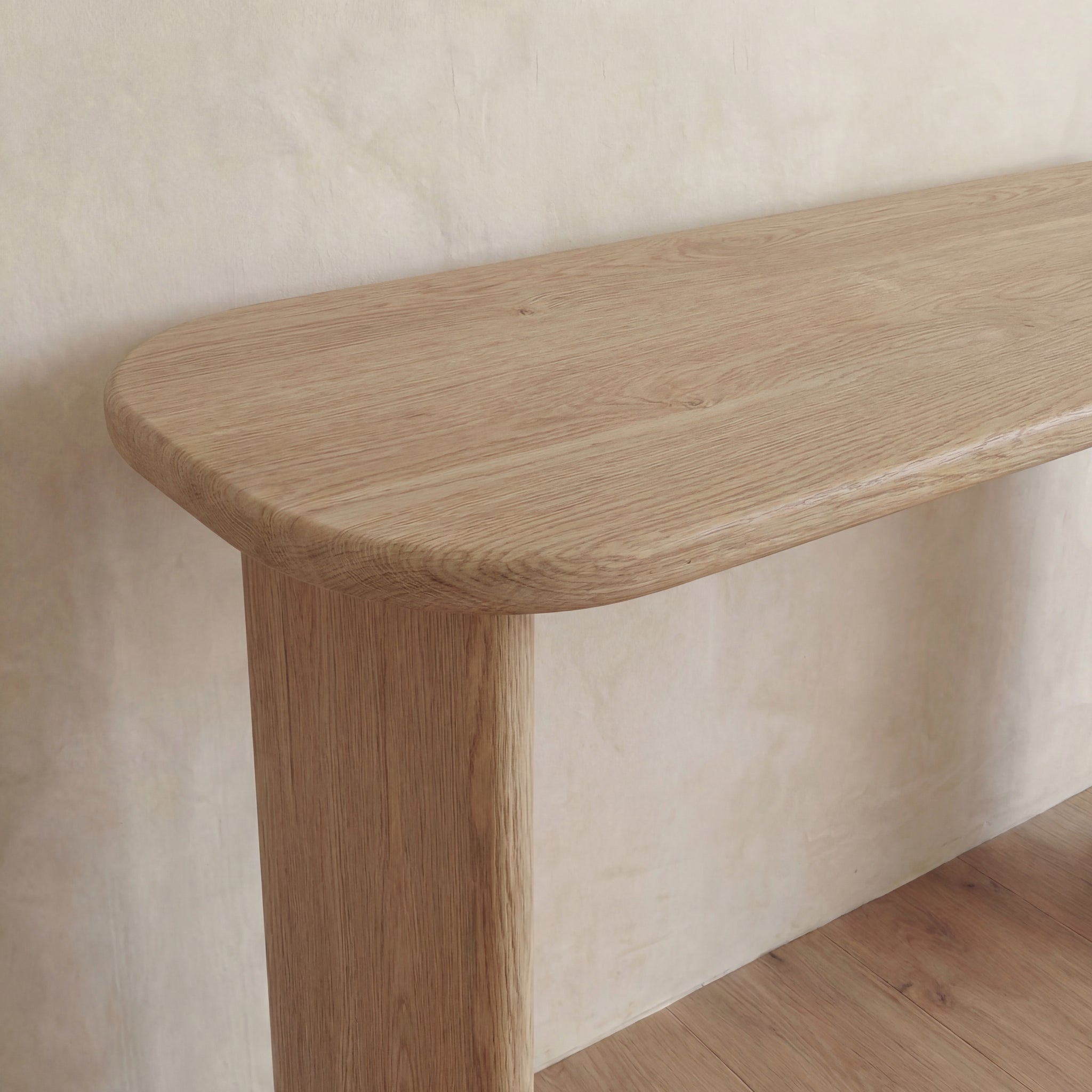 A close-up of the 60 inch console table in white oak, featuring rounded corners and a light natural finish, set against a textured beige wall on a wooden floor.