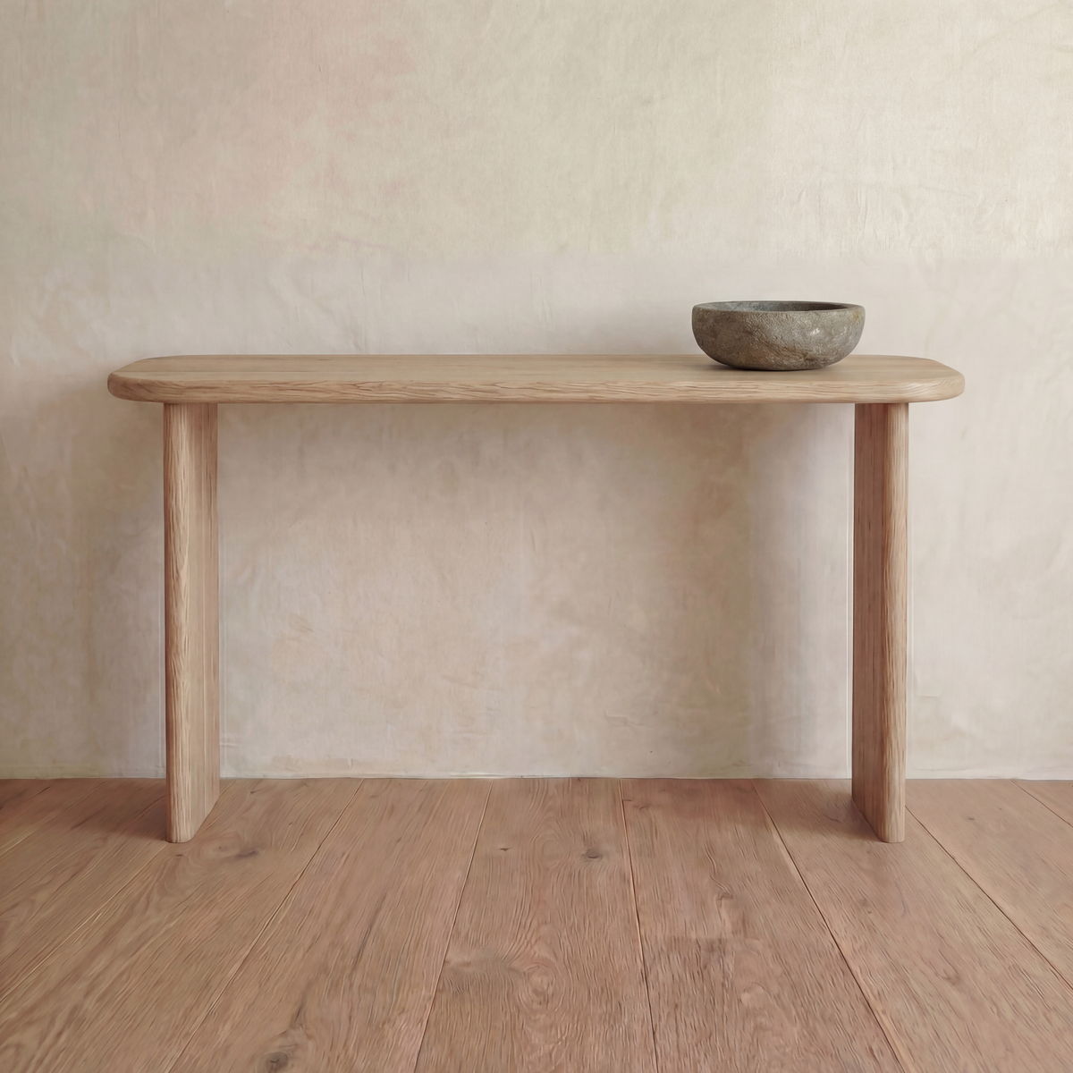 A 60 inch console table in white oak with rounded edges and two legs, with a stone bowl on top, stands against a textured beige wall on a wooden floor.