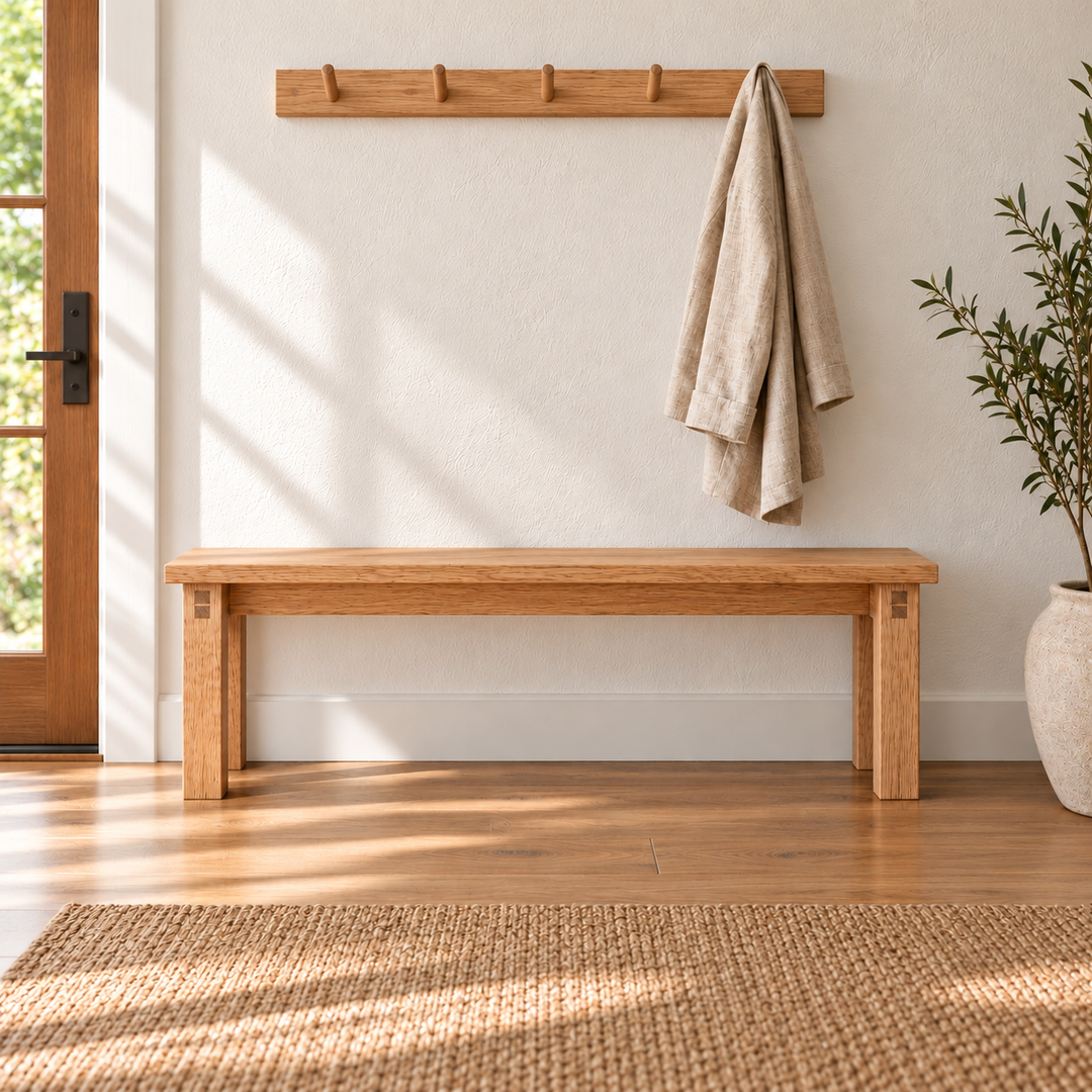 A farmhouse bench in red oak in a sunlit entryway, below a wooden peg rail with a linen towel, beside a potted olive tree and a woven rug.